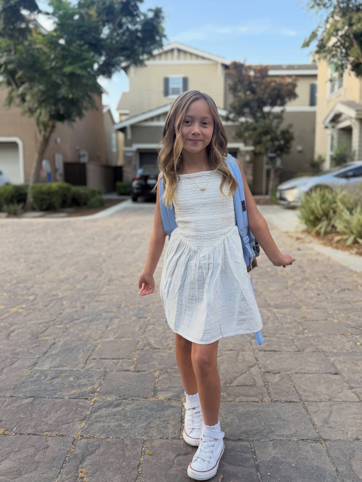 Picture Day Outfits That Photograph Beautifully (7 Ideas) 6 Young girl in a white dress and blue backpack standing on a paved driveway.
