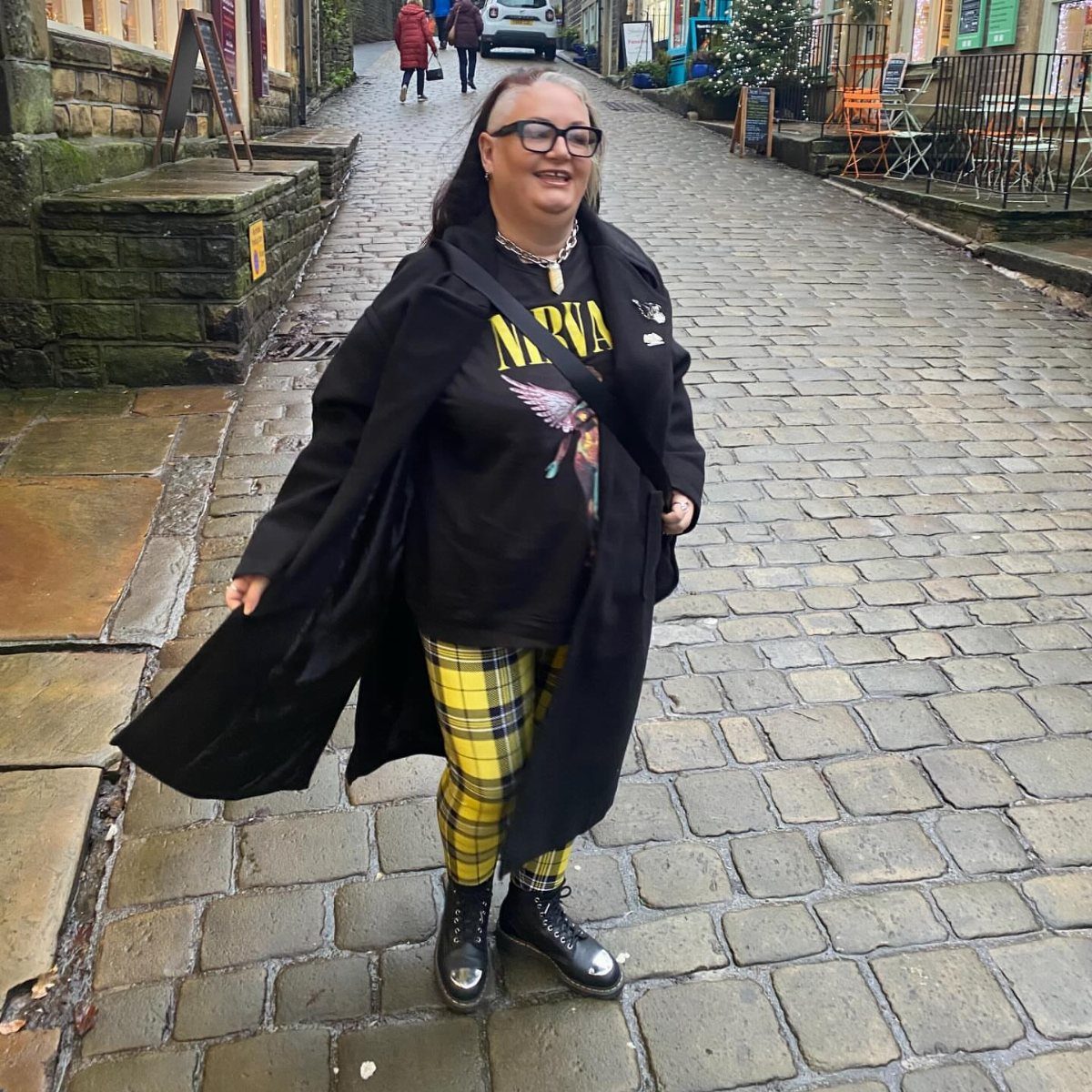 Plus Size Winter Outfits: 7 Cozy and Stylish Looks to Try 2 Plus-size woman in a long black coat and yellow plaid pants on cobblestones.