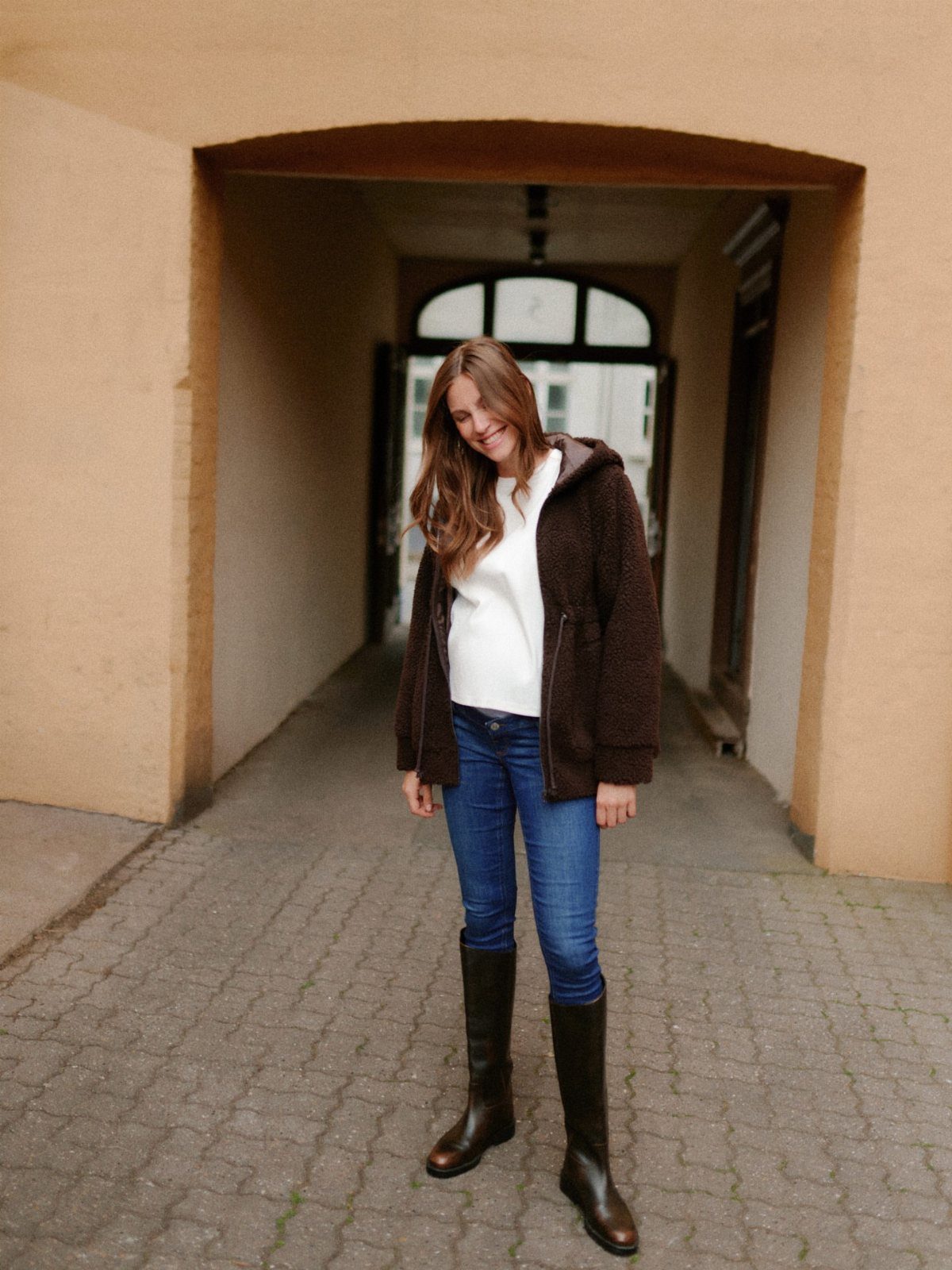 12 Pregnant Fall Outfits That Work Through Every Trimester 4 Pregnant woman wearing a brown fleece jacket, white top, blue jeans, and brown boots.