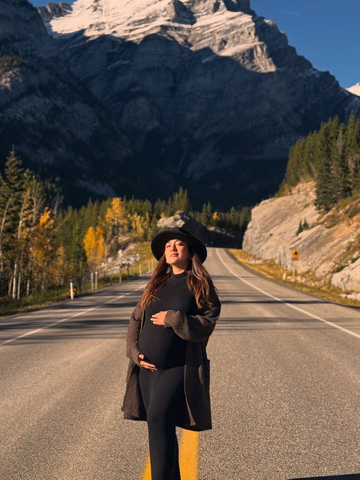 12 Pregnant Fall Outfits That Work Through Every Trimester 2 Pregnant woman in a long brown cardigan and black outfit on a mountain road