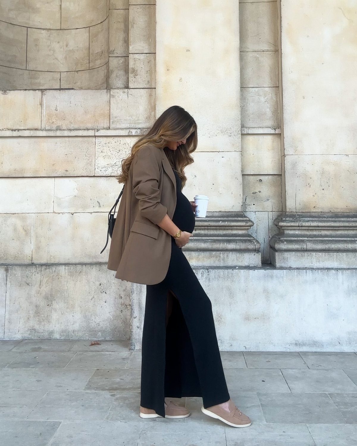 12 Pregnant Fall Outfits That Work Through Every Trimester 7 Pregnant woman in a brown blazer and black wide-leg trousers standing by stone wall.
