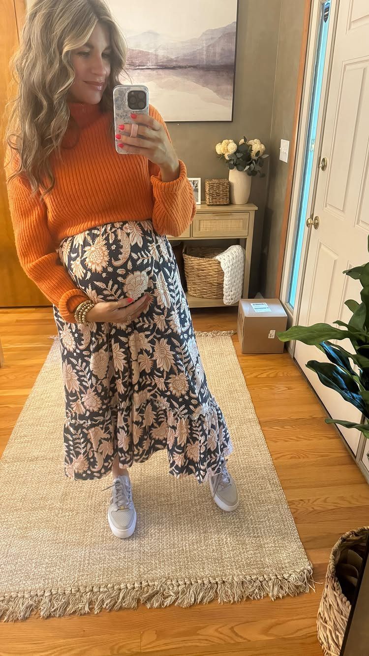 12 Pregnant Fall Outfits That Work Through Every Trimester 10 Pregnant woman wearing an orange sweater and long floral skirt with white sneakers.