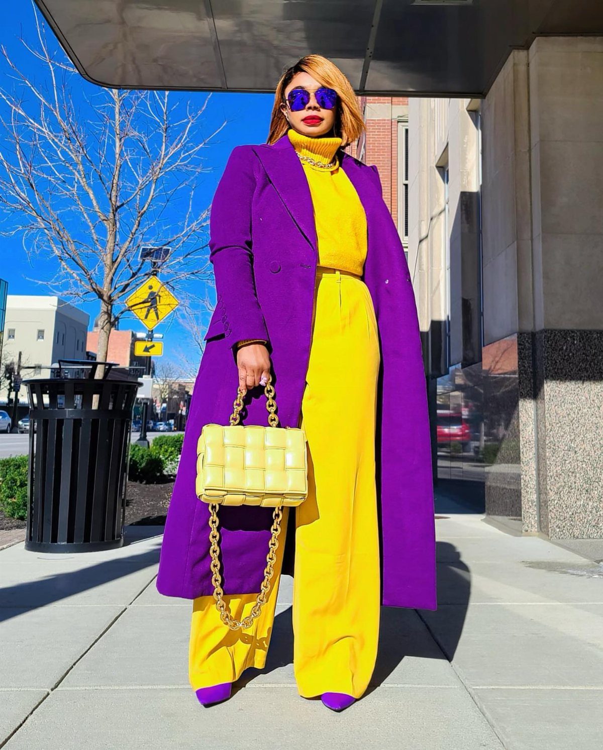 Purple Winter Outfits: 9 Ways to Style the Season's Boldest Color 8 Woman in a long purple coat and yellow outfit standing on a city sidewalk.