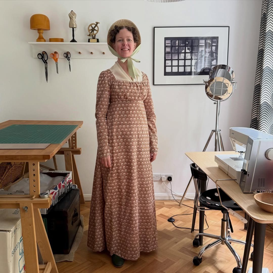 Regency Era Outfits Women Wore: 9 Elegant Period Pieces 1 Woman in a brown empire waist Regency dress and straw bonnet in a studio.