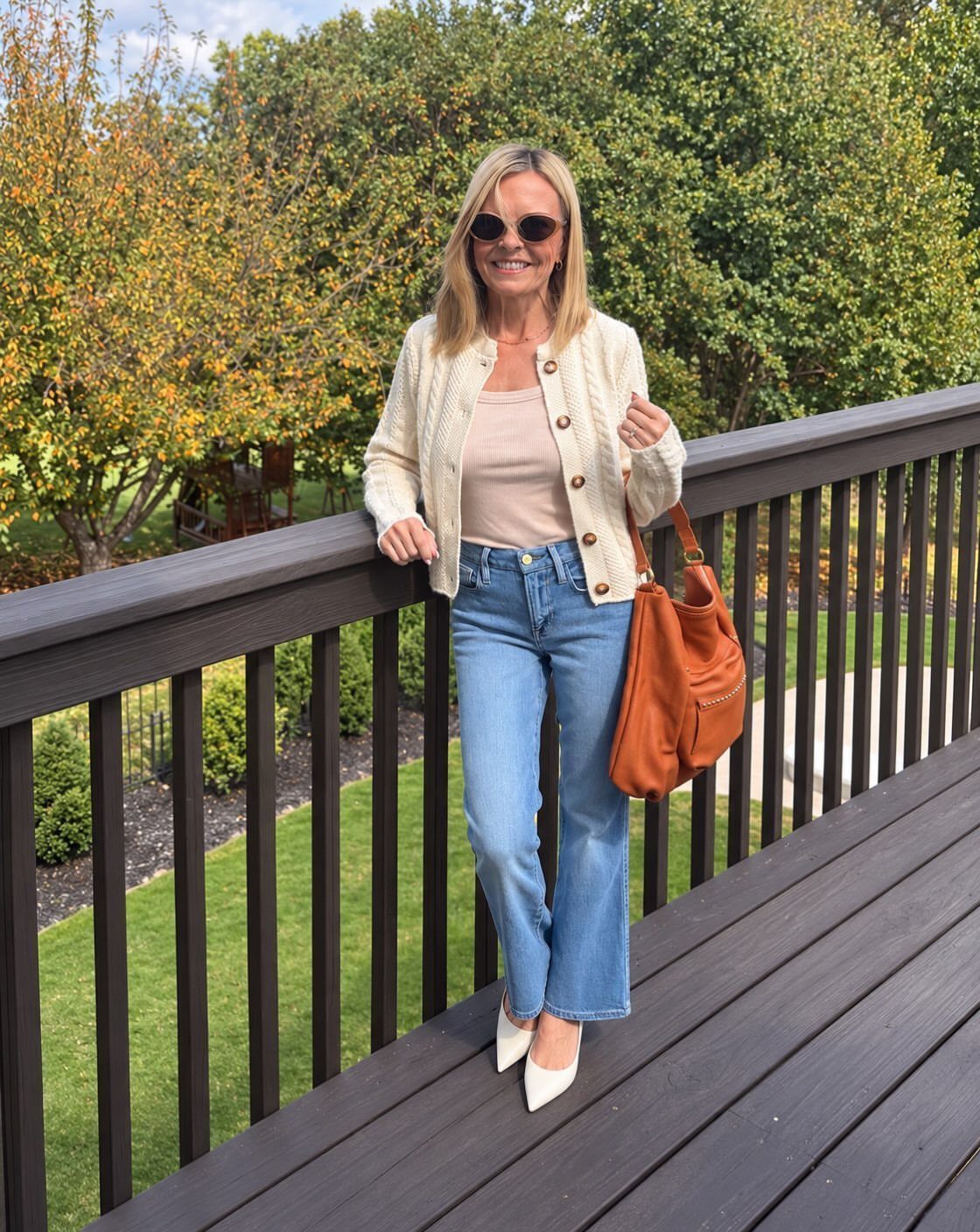 16 Short Torso Outfits That Balance Your Proportions 12 Woman in cream cardigan, beige tank, and blue mid-rise jeans on a wooden deck.