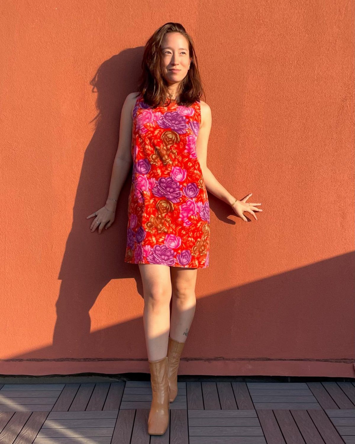 16 Short Torso Outfits That Balance Your Proportions 3 Woman in a floral shift dress and tan boots posing against an orange wall.