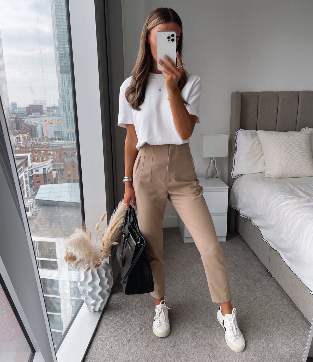 12 Spring Work Outfits That Work From Desk to Dinner 1 Woman wearing tan tailored trousers, a white t-shirt, and white sneakers in a bedroom.