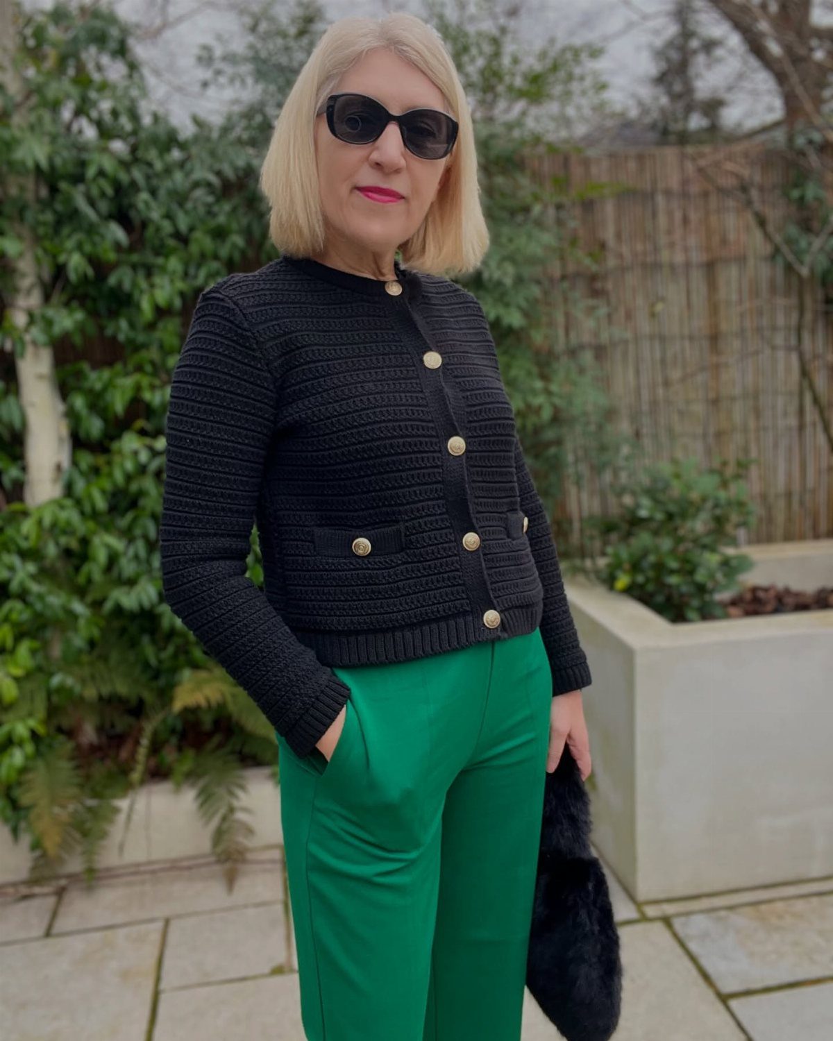 12 Spring Work Outfits That Work From Desk to Dinner 11 Woman wearing a black buttoned cardigan and bright green trousers standing outdoors.