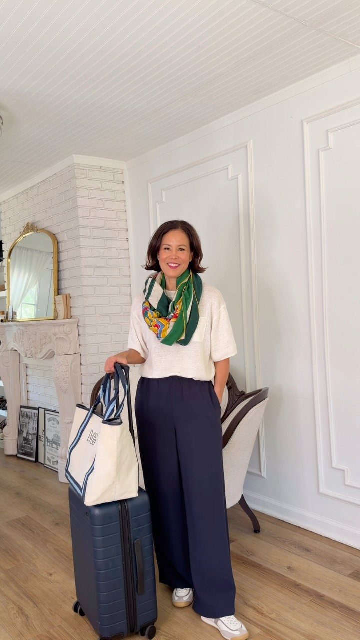 Woman wearing white knit top and navy trousers standing with a blue suitcase.