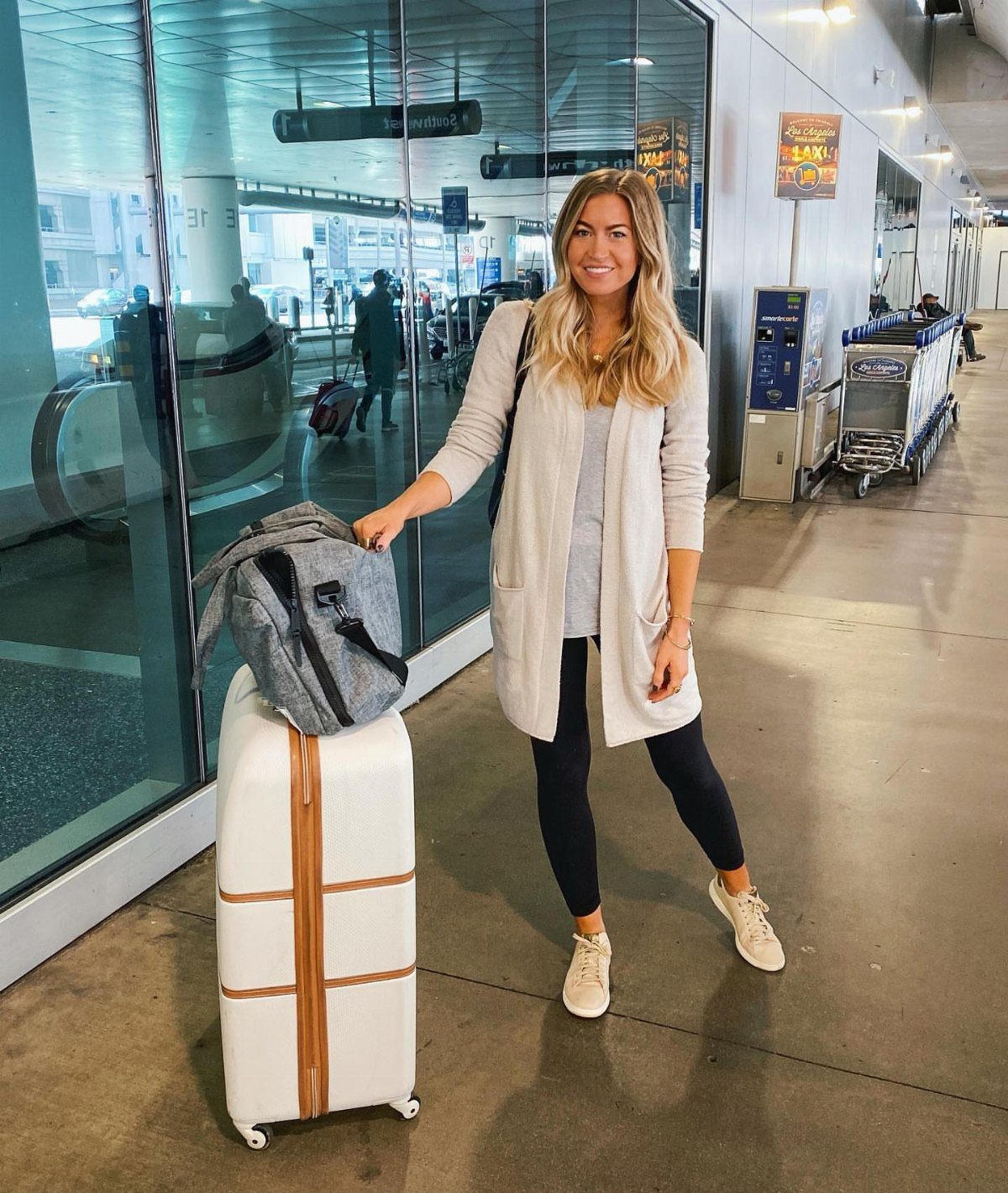 Woman in beige long cardigan and black leggings standing with white suitcase in airport