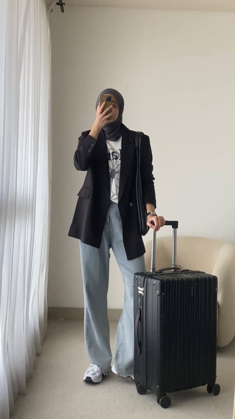 Person in black blazer and light blue jeans standing with a black suitcase.