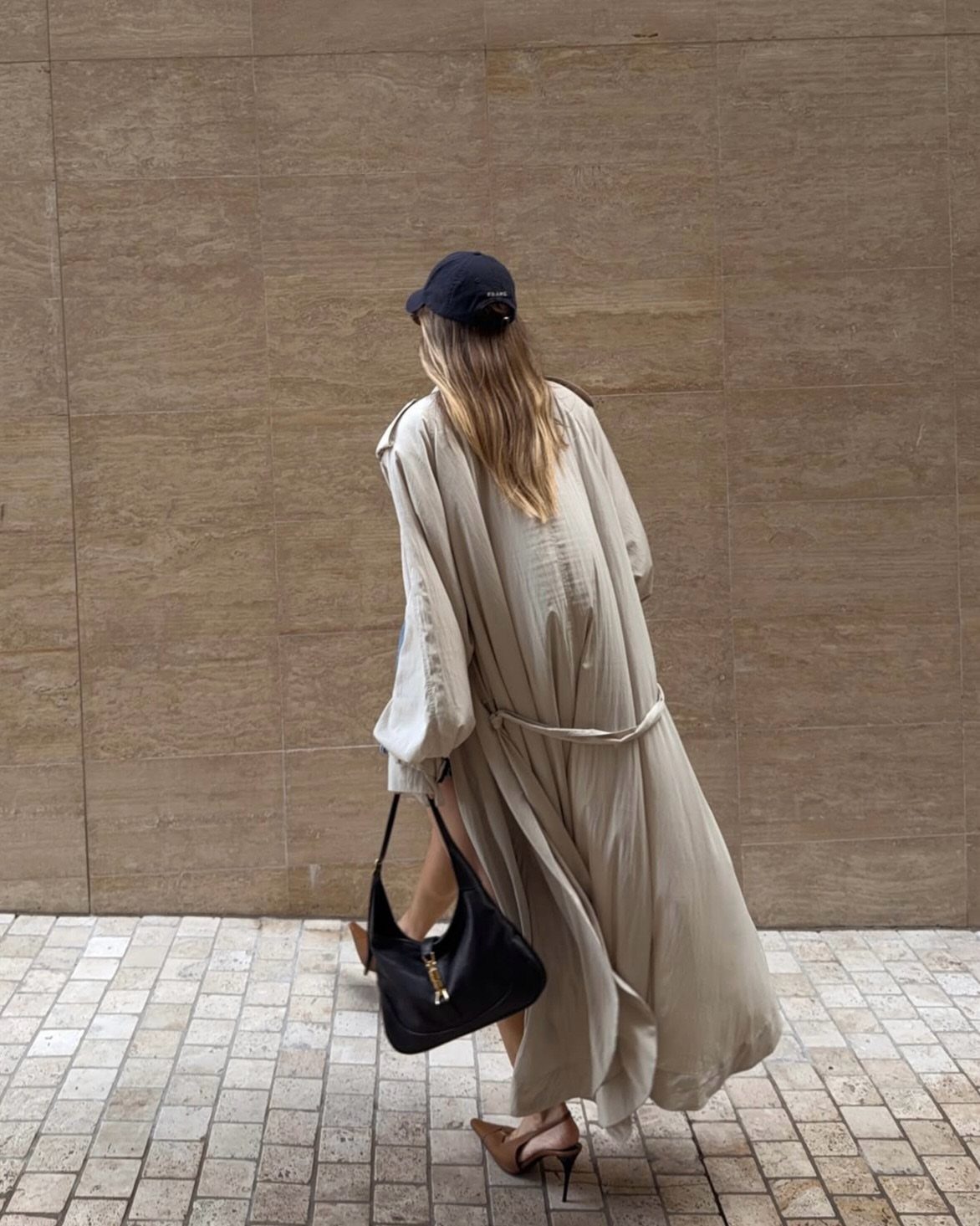 Woman wearing a long fluid beige trench coat, black cap, and black handbag.