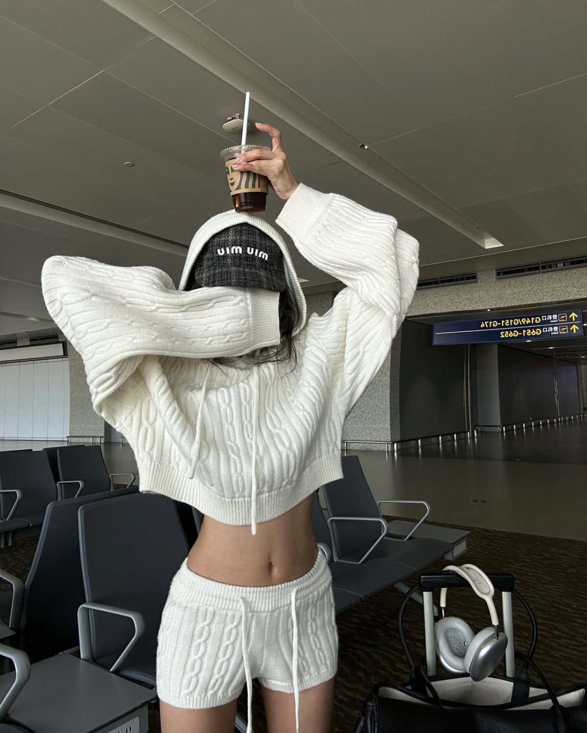 Person wearing a white cable-knit cropped pullover and matching shorts in an airport terminal.