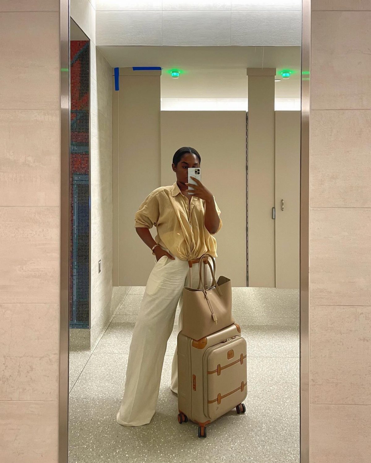 Woman in yellow shirt and white linen pants posing with luggage in airport.