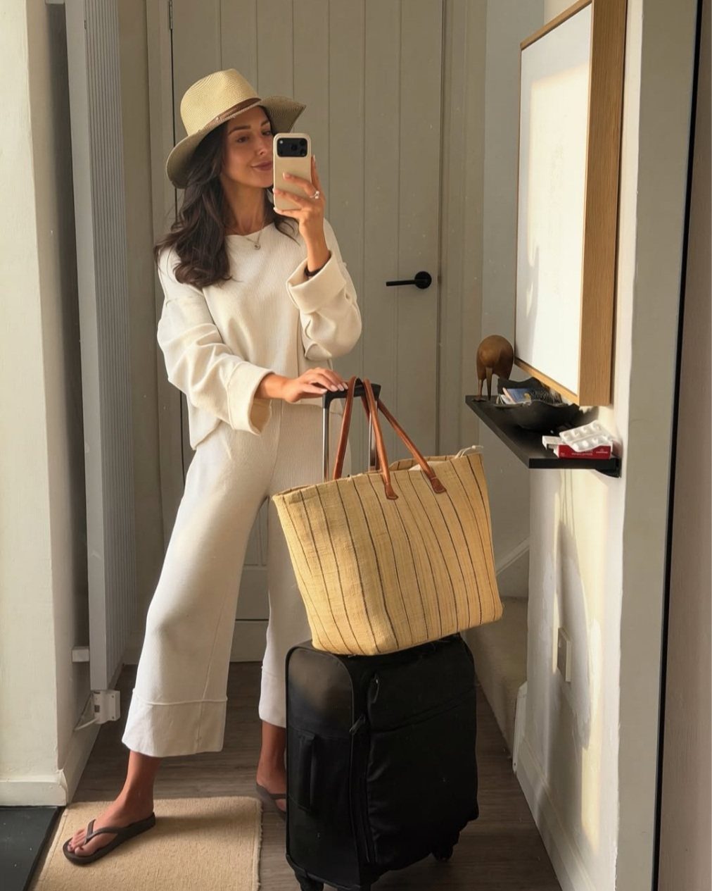 Woman in white outfit and tan hat standing with suitcase and straw tote bag.