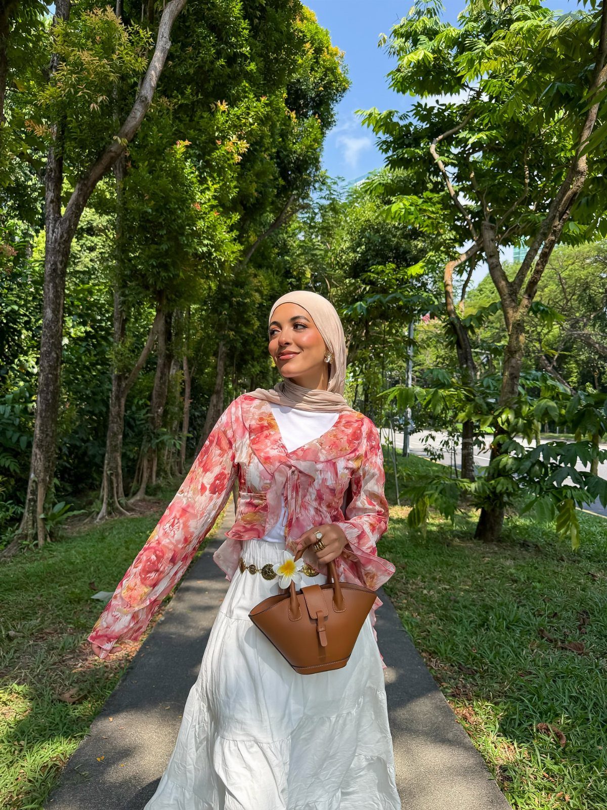 8 Summer Hijab Outfits for Modest, Breathable Style 8 Woman in a floral blouse, white skirt, and beige hijab posing in a park.