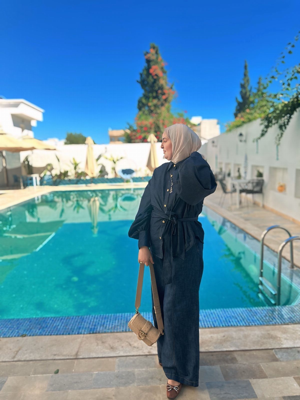 8 Summer Hijab Outfits for Modest, Breathable Style 4 Woman in a black jumpsuit and beige hijab standing beside a blue swimming pool.