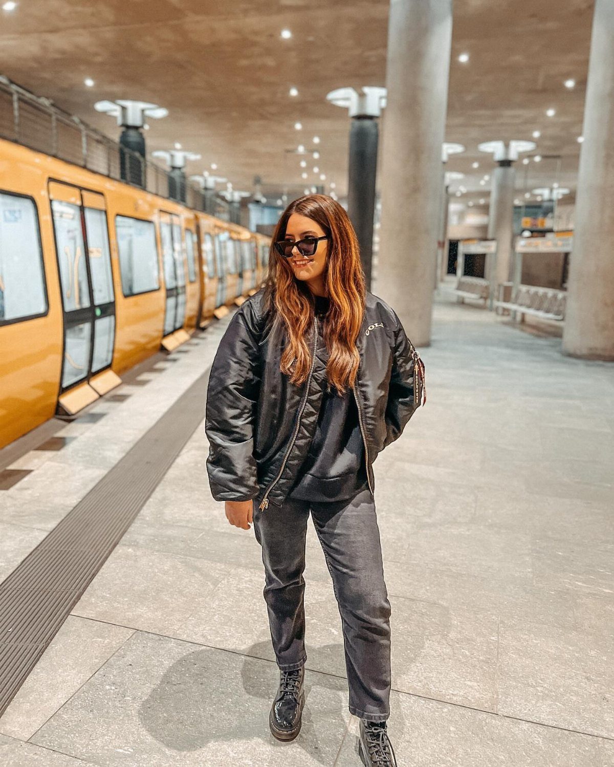 12 Super Bowl Outfits From Sporty to Sequined for Game Day 4 Woman wearing a black bomber jacket and grey jeans on a train platform.