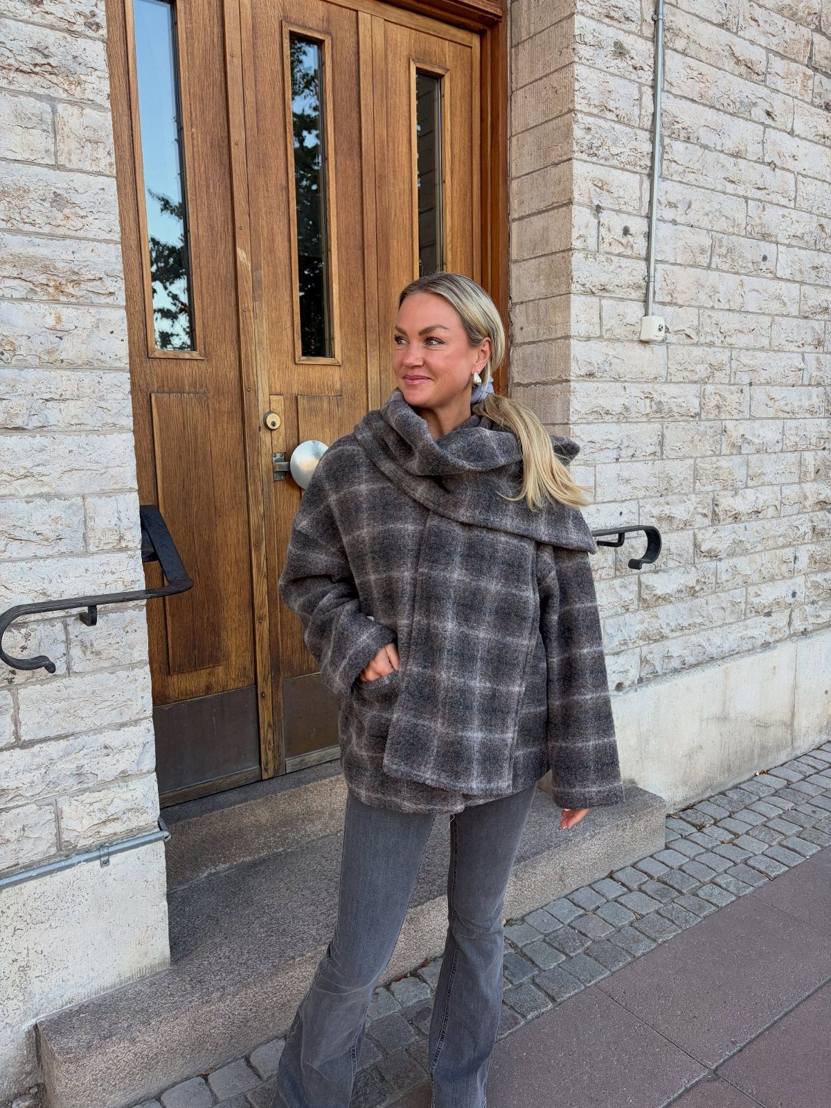 Women's Scarf Coat: 12 Ideas to Try This Season 11 Woman wearing a grey plaid scarf coat and grey trousers by a wooden door.