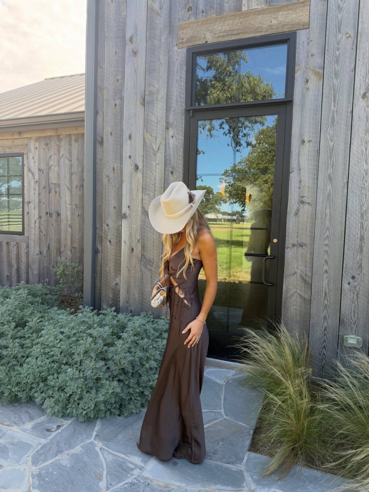 Women's Wild West Outfit Ideas: 9 Styles That Deliver 8 Woman in a brown maxi dress and white cowboy hat standing by wooden building