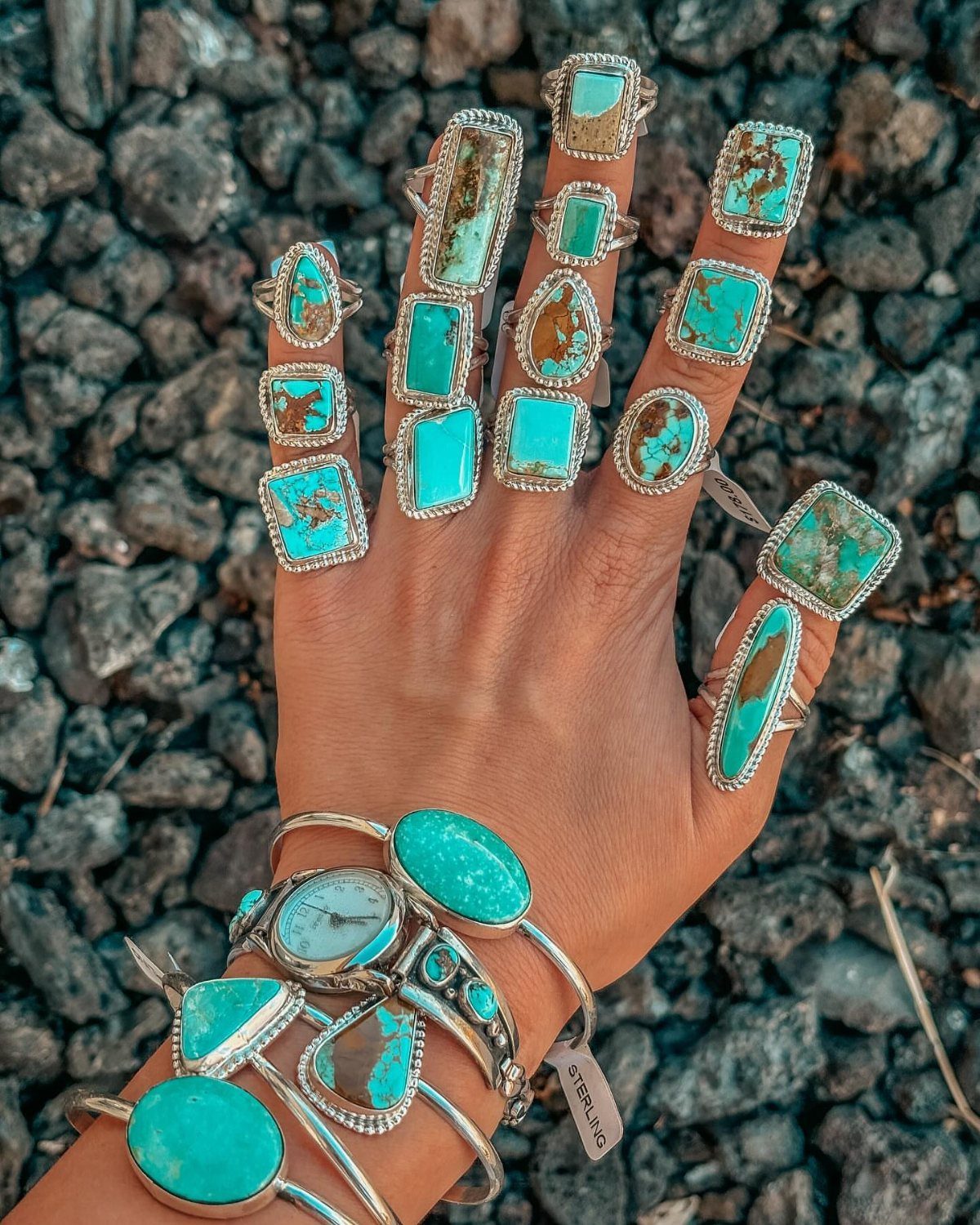 Women's Wild West Outfit Ideas: 9 Styles That Deliver 9 Hand and wrist wearing multiple silver turquoise rings and bracelets over dark pebbles.
